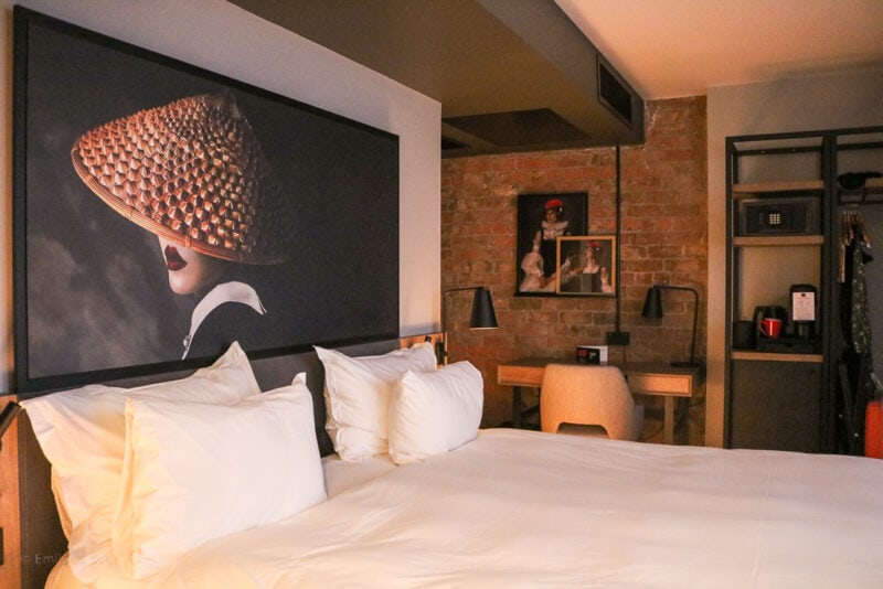 Hotel room at the Readisson RED in Liverpool with an exposed brick wall and a large double bed with white sheets. Over the bed is a large painting of a giesha with her face mostly covered by a straw hat on a black background.