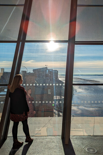 Emily wearing black tights and a long black cardigan standing in front of a large glass window with a view of a river and the liverpool docks below