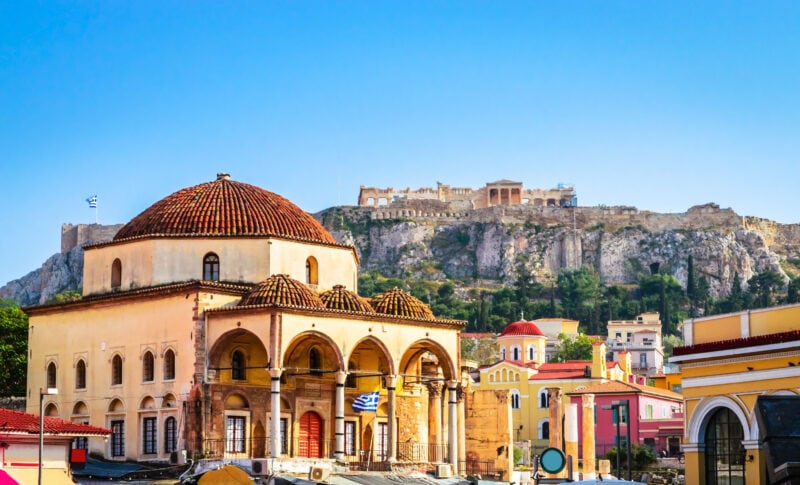 Large Ottoman mosque in Monastiraki square with yellow walls and a red tirled dowed roof, there are collonaded arches around the front section on the ground floor. Behind the church is a small rocky hill with the ancient ruins of the acropolis on top. unusual things to do in athens. 