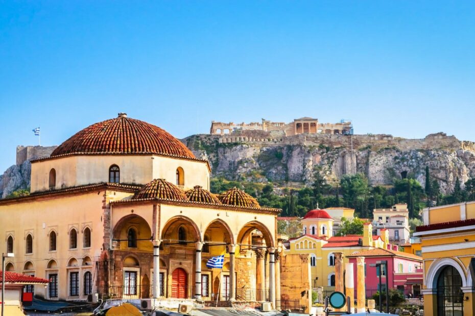 Large Ottoman mosque in Monastiraki square with yellow walls and a red tirled dowed roof, there are collonaded arches around the front section on the ground floor. Behind the church is a small rocky hill with the ancient ruins of the acropolis on top. unusual things to do in athens.