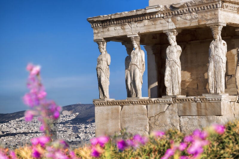Side of the Parthenon temple with four marble sculptures of human figures that form columns supporting the roof, taken during spring time on the Acropolis in Athens with bright pink flowers out of focus in the foreground and clear blue sky behind