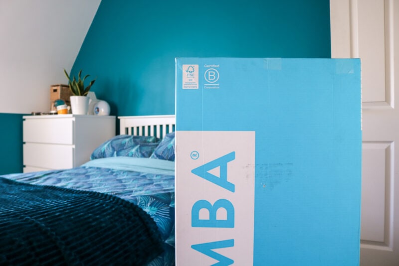 cyan and white cardboard box with the letters M B A on the side in front of a double bed with turquoise sheets and a white wooden headboard with a turquoise painted wall behind