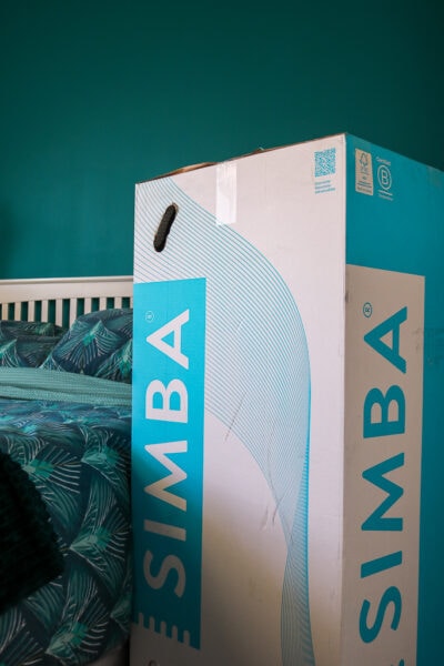 cyan and white cardboard box in front of a double bed with turquoise sheets and a white wooden headboard with a turquoise painted wall behind. The box has the word SIMBA printed on both sides.