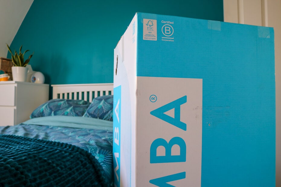 cyan and white cardboard box with the letters M B A on the side in front of a double bed with turquoise sheets and a white wooden headboard with a turquoise painted wall behind