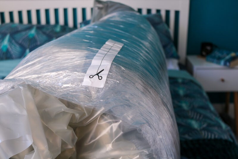 Close up of a roll of plastic with a vacuum-packed mattress inside on a bed with turquoise sheets