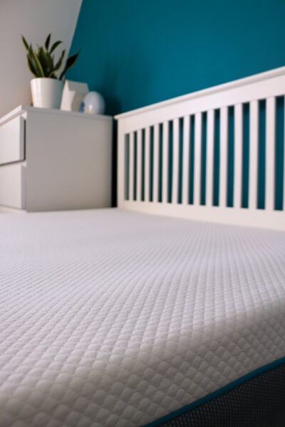 Close up of part of a white mattress with a soft patterned top on a white wooden bedframe next to a white chest of drawers with a turquoise wall behind. simba Hybrid mattress review