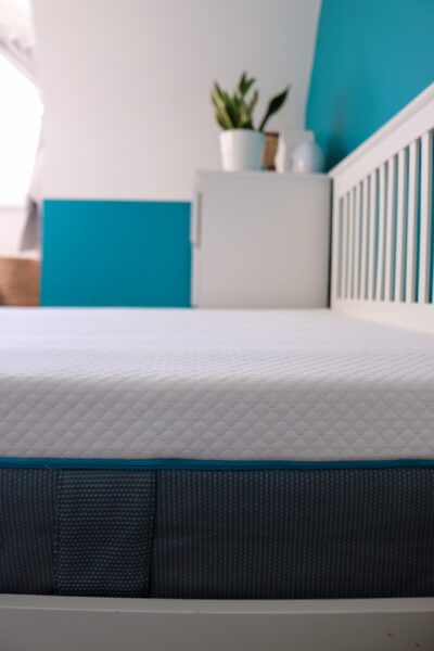 Close up of part of a white mattress with a soft patterned top and a grey base on a white wooden bedframe next to a white chest of drawers with a turquoise wall behind