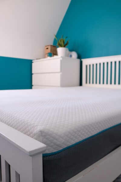 Close up of part of a white mattress with a soft patterned top and a grey base on a white wooden bedframe next to a white chest of drawers with a turquoise wall behind. Simba Hybrid Original Mattress Review.