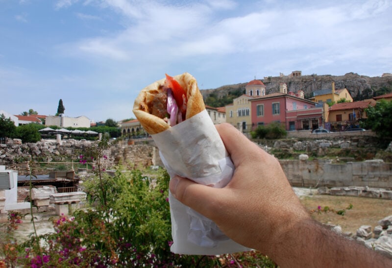 man's hand holding a souvlaki or greek kebab, with meat wrapped in pita in a white paper wrap, in front of a view of the athens skyline