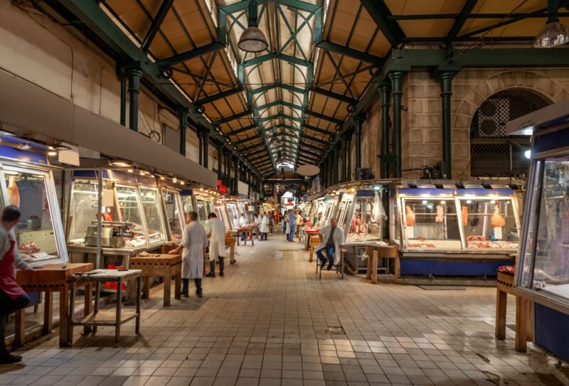 Interior of Varvakios Central Market in Athens, a large hall with high cieling with green metal beams. On either side is a row of stalls with lit up glass fronts and vendors wearing white coats
