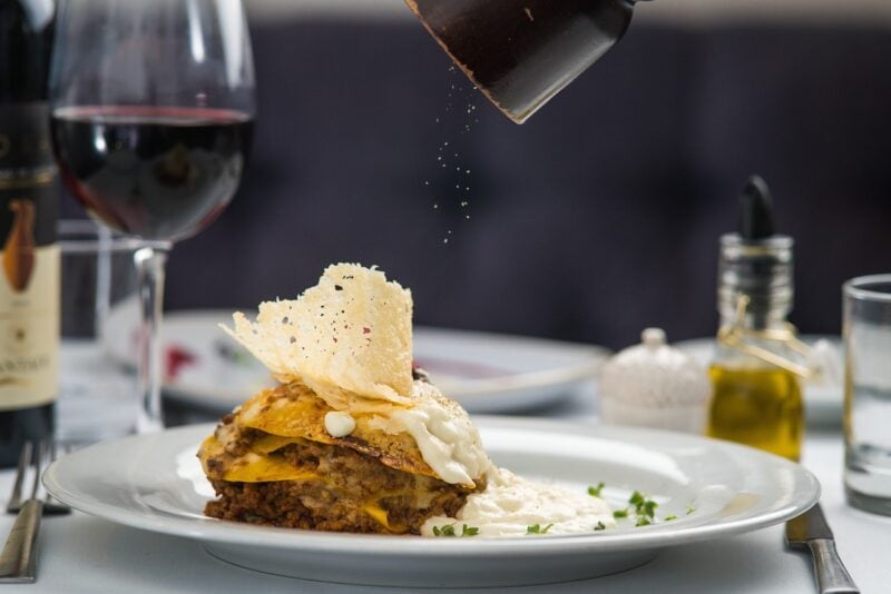 small piece of lasagna on a white plate next to a glass of red wine