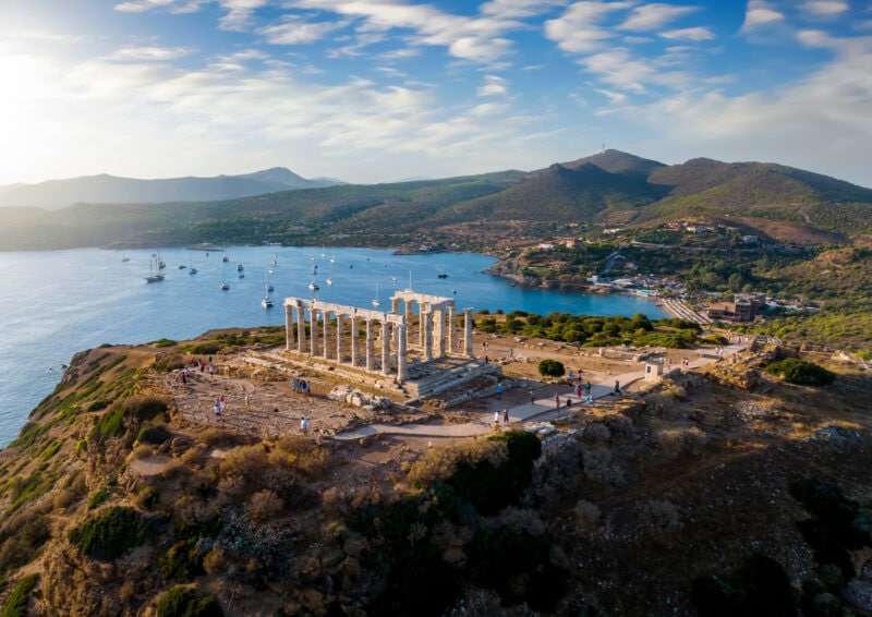 Aerial view of the a small bay with a beach and Temple of Poseidon at Cape Sounion at the edge of Attica, Greece, during summer sunset time