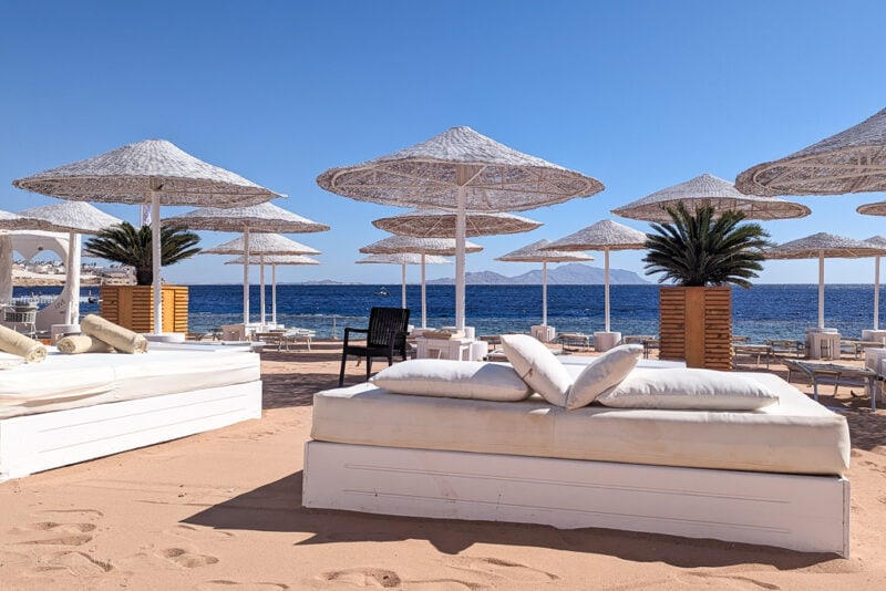 white double sunbed on a sandy beach with rows of pale wooden umbrellas behind and the sea behind that on a very sunny day with clear blue sky