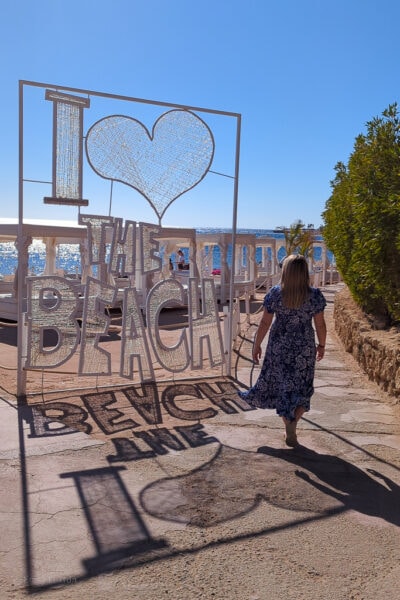 emily wearing a long blue dress with white swirls walking away from the camera towards a sandy beach next to a large white sign that says I heart the beach, taken on a sunny day with clear blue sky
