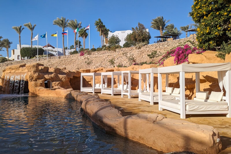 outdoor swimming pool surrounded by an orange rocky wall with a small waterfall at the far end and a row of white four poster sunbeds along the right hand side. Elisir Spa at domina coral bay sharm el sheikh