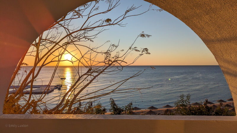 large archway in a white wall with a view of the sun rising over the blue sea, there is a bare tree in the foreground with a silhouette of a bird in the branches. 