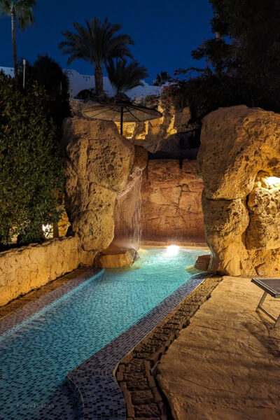 small, shallow pool with a rocky cave at one end with a waterfall flowing down one side, taken at night and lit with orange lights