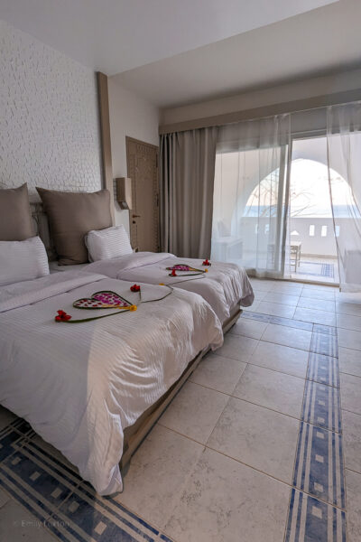 hotel room with white walls and pale grey tiled floors with a blue line pattern in a large square around the room. There are two single beds with white bedding and large grey pillows, and flowers in the shape of a heart on the beds. Aquamarine beachfront room at domina coral bay sharm el sheikh