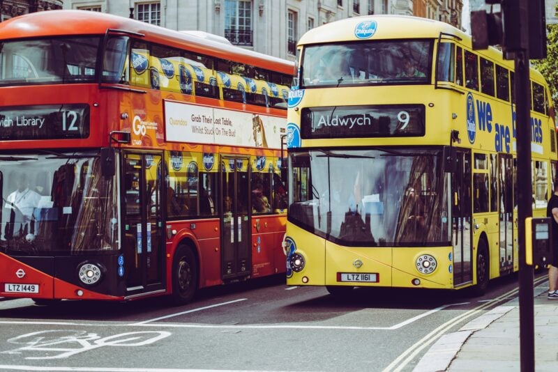 a red double decker bus next to a yellow double decker bus on a grey tarmac road.