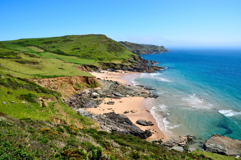 long row of grassy cliffs on the south devon coast with two sandy beaches below in between large grey rocks with the bright blue sea on the right and blue sky above