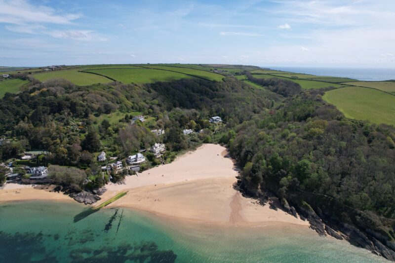 aerial view of a small cove with a white sandy beach next to a river with turquoise water surrounded by green grassy hills and woodland