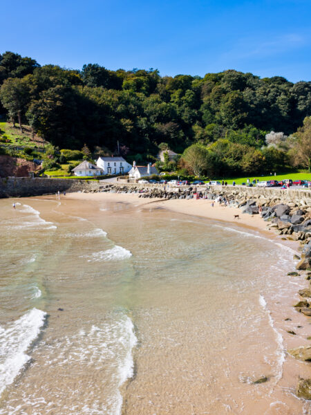 small cove with a sandy beach and grey rocks at high tide with shallow waves on the sand and a hill on the far side of the cove covered with green trees on a sunny day