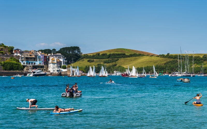 a man and woman on blue paddleboards on a wide blue river with a motorboat behind them and many white sailing boats in the distance