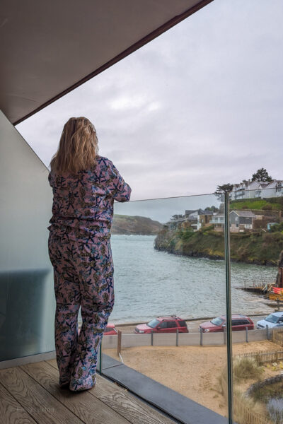emily wearing pink pyjamas with a blue pattern on standing on a balcony next to a glass wall looking out to sea with a sandy beach below and grey sky above at South Sands Beach in Salcombe