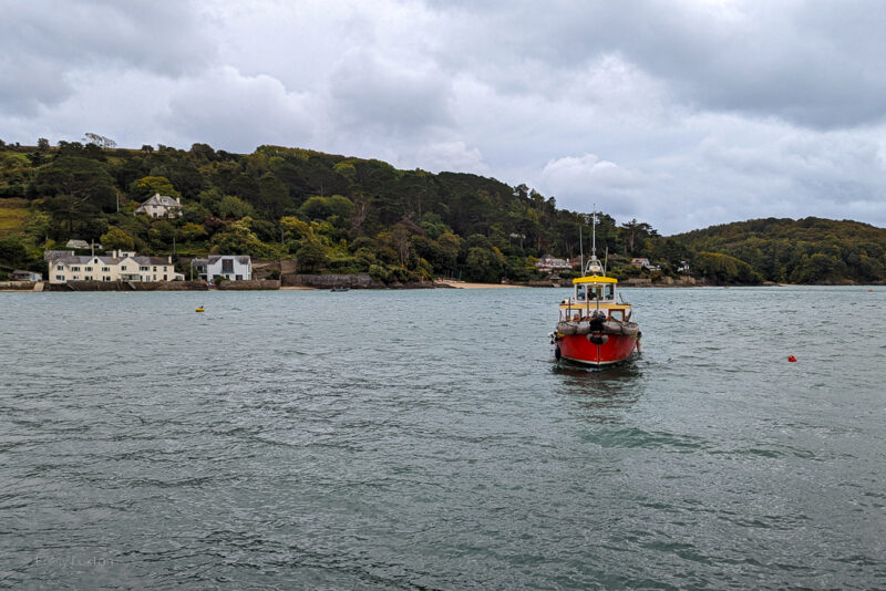 red, white and yellow ferry boat on a river with a green hill behind on a grey day with cloudy sky - the Kingsbridge to Salcombe ferry heading towards the jetty
