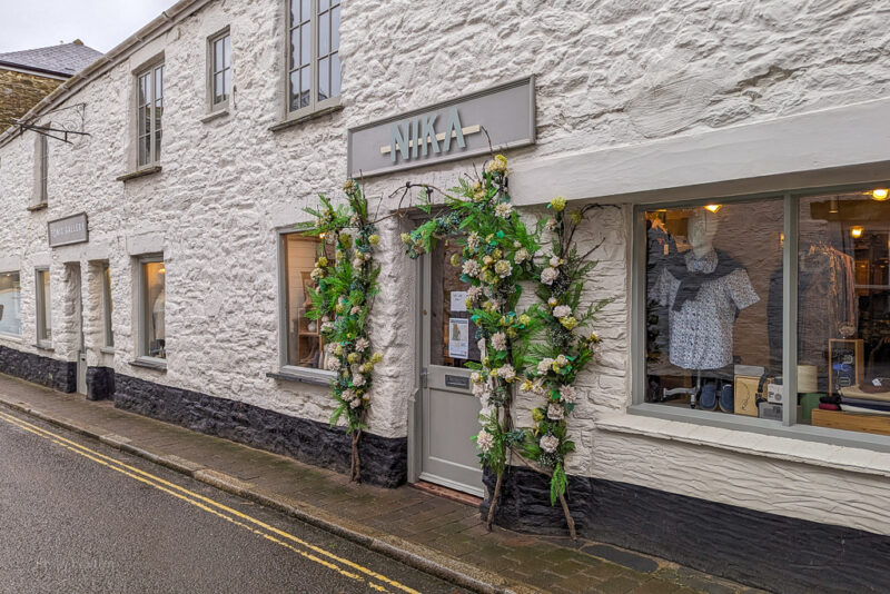 whitewashed stone cottage with a shop front, there are green and pink flowers growing around the grey front door and the sign above says NIKA