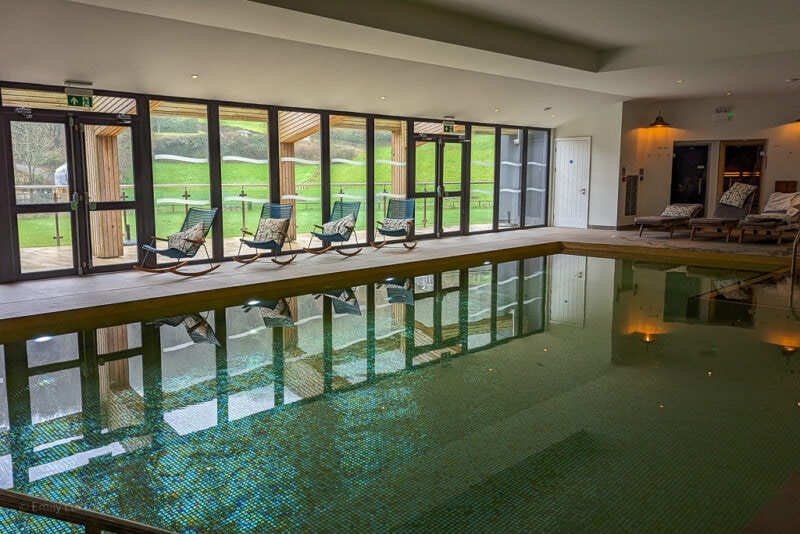 rectangular indoor swimming pool with four beach chairs lined up next to it infront of large windows overlooking a grassy field