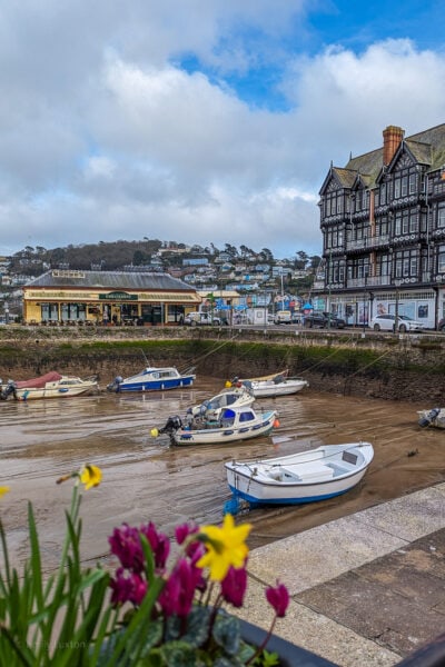 view of a dry harbour with several small boats moored in the mud. there is a white and black timber clad tudor building behind and pink flowers and daffodils in the foreground.