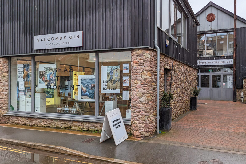 exterior of Salcombe Gin Distillery with brick walls on the lower half and grey wood cladding on the top hald. There is a large window with bottles of gin and posters on sdisplay.