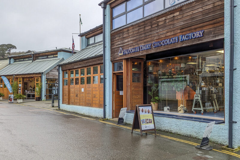 exterior of Salcombe Dairy Chcocolate Factory, a blue painted building with wooden cladding along the top floor. there is a large window with chocolate making instruments visible.