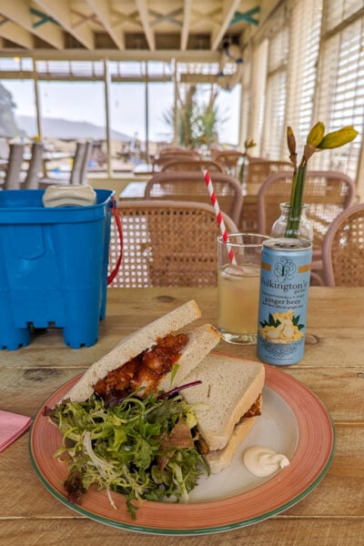 white bread sandwich filled with breaded shrimp next to a green leaf salad on a white and red plate on a wooden table in a restaurant with cream walls and a large window. The Winking Prawn Restaurant in Salcombe. 