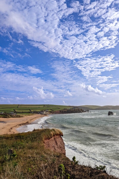 sandy beach with grassy hills behind on a windy, sunny day with rough waves and blue sky