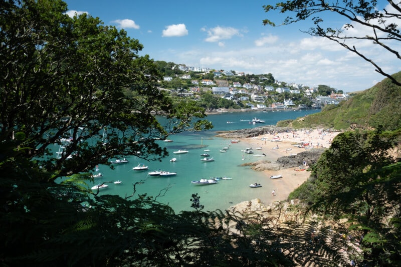 view between trees of a river with a white sandy beach on the right bank and a hill with a small town on the far side on a sunny day with turquoise water and blue sky. Sunny Cove on Kingsbridge Estuary, one of the best beaches in Salcombe. 