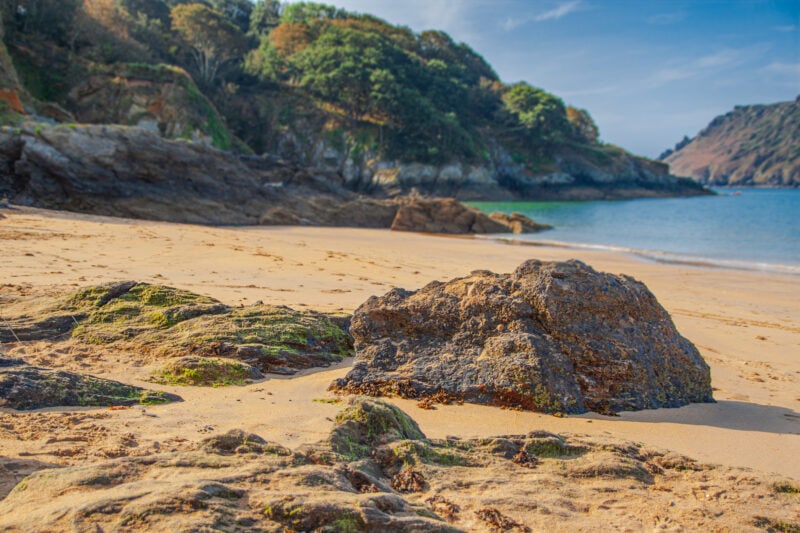 low view of a rock on a golden sandy beach with hills and blue sea in the background