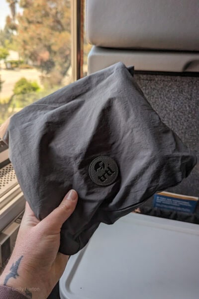 main tenant un petit sac en tissu gris avec un logo circulaire indiquant TRTL devant une fenêtre de train avec des arbres flous à l'extérieur