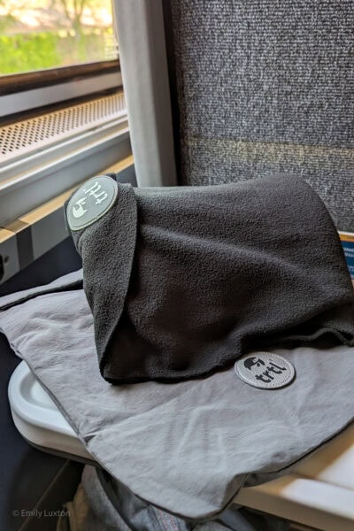 carré plié en polaire noire avec un logo gris indiquant TRTL.  La polaire est posée sur un sac gris portant le même logo, sur une table rabattable dans un train avec des arbres flous visibles par la fenêtre.