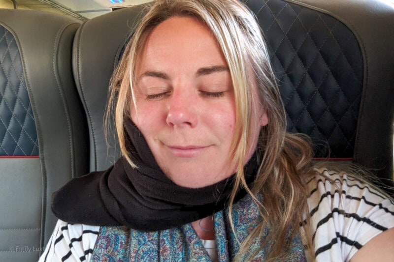 Selfie d'Emily, les yeux fermés, sur un siège de train en cuir bleu, portant un t-shirt blanc à rayures noires et une écharpe en polaire noire enroulée autour de son cou