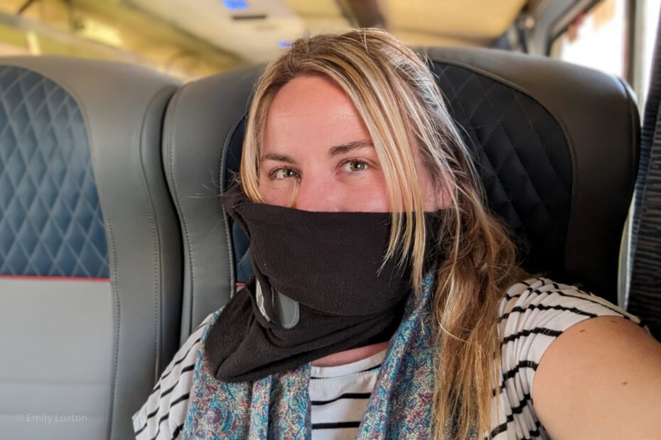 Selfie of Emily on a blue leather train seat wearing a white t shirt with black stripes and a black fleece scarf wrapped around her neck - the TRTL Travel Pillow