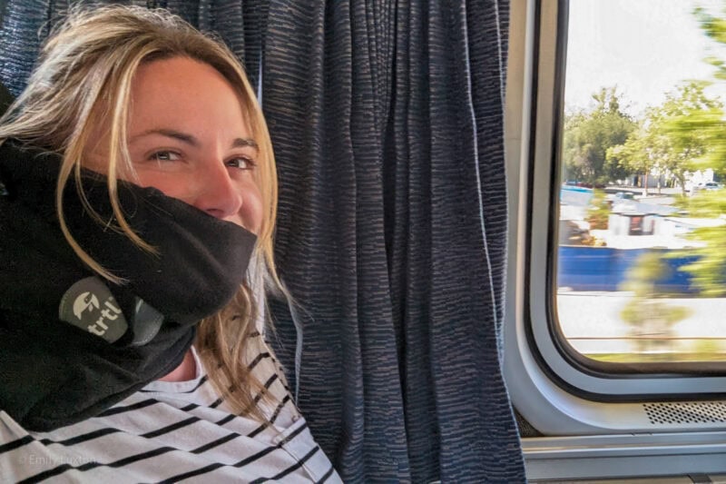 Selfie d'Emily sur un siège de train en cuir bleu portant un t-shirt blanc à rayures noires et une écharpe en polaire noire enroulée autour de son cou - TRTL Travel Pillow Review