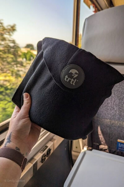 main tenant l'oreiller de voyage TRTL devant une fenêtre de train avec des arbres et un ciel bleu à l'extérieur, l'oreiller est un carré de tissu polaire plié avec un cercle gris contenant le logo TRTL.