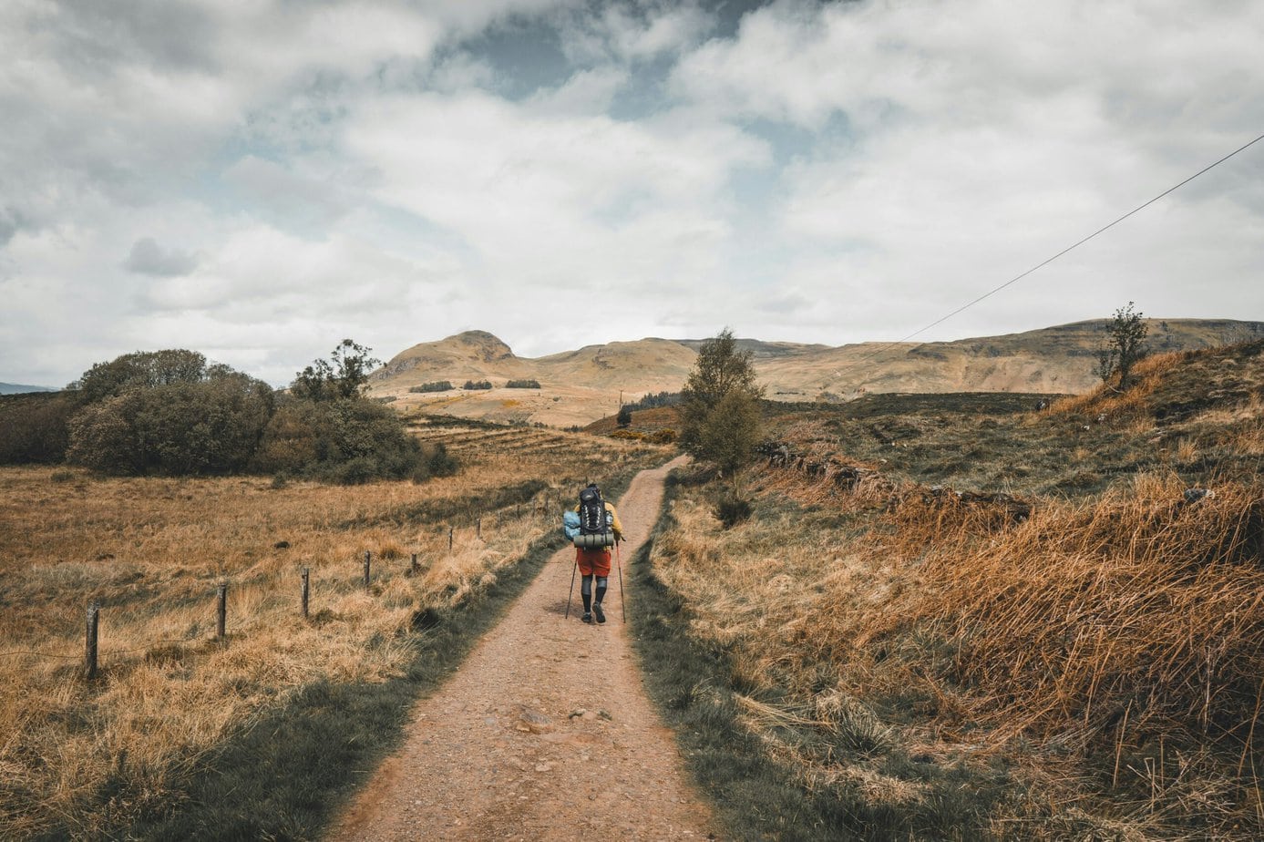 person in red jacket walking on brown dirt road in day time with brownish grassy hills beyond