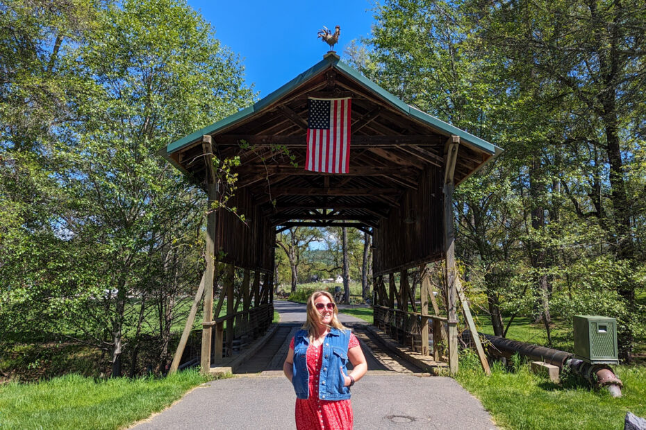 Emily standing with her hands in her pockets in front of a wooden covered bridge with a US flag hanging from the top of it and trees on either side. Emily is wearing a long red dress and a blue denim waistcoat and has her long hair loose.