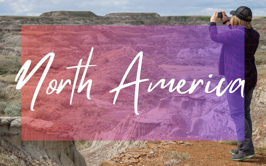 North America written in white text over an opaque pink box on top of a photo of an arid landscape with a rocky valley and beige coloured sandy hills. Travel blogger Emily Luxton is standing on a cliff edge in the foreground holding her camera up to take a photo. She is wearing a dark red checked flannel shirt and grey trousers.