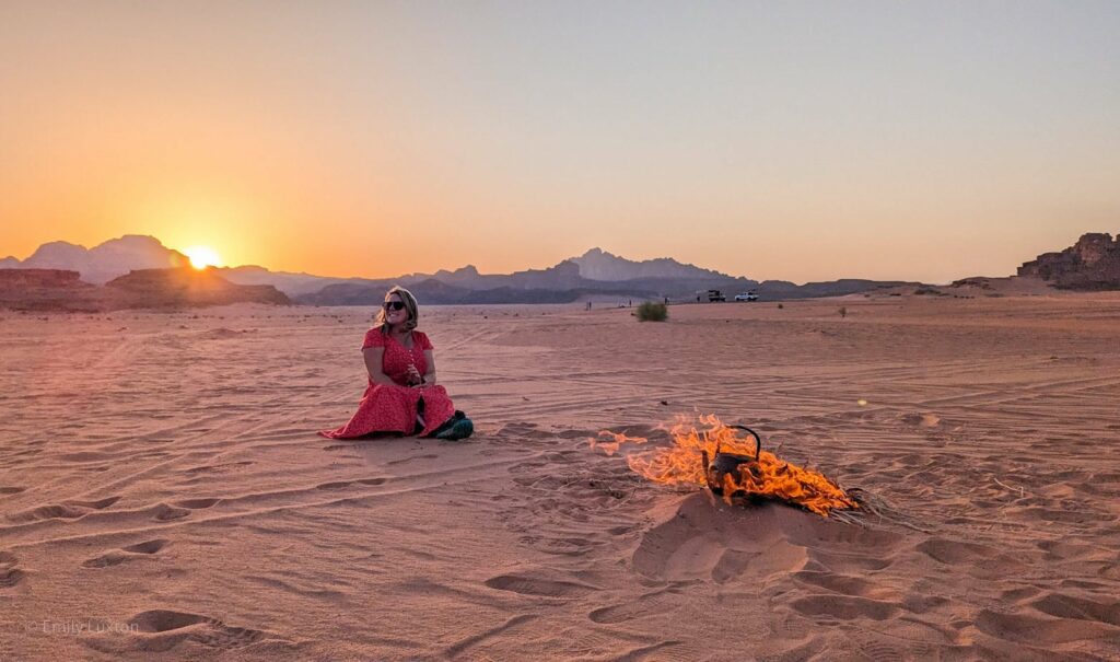 empty desert landscape with the sun setting behind low mountains in the distance. There is a campfire with an iron kettle in the flames and Emily is sat in the sand behind it wearing a red dress.