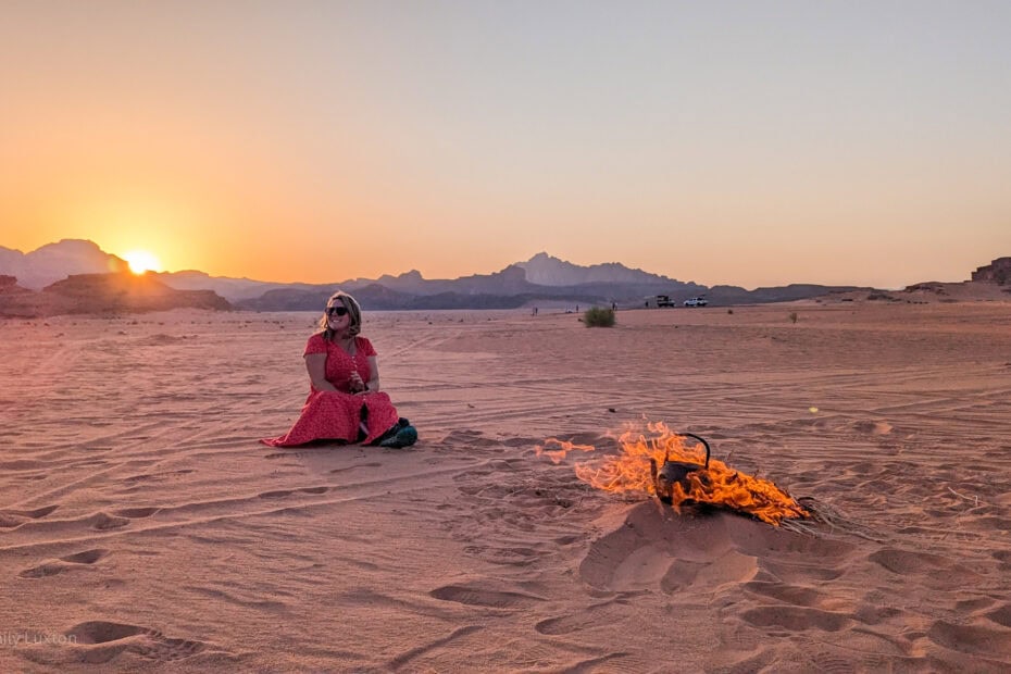 empty desert landscape with the sun setting behind low mountains in the distance. There is a campfire with an iron kettle in the flames and Emily is sat in the sand behind it wearing a red dress.