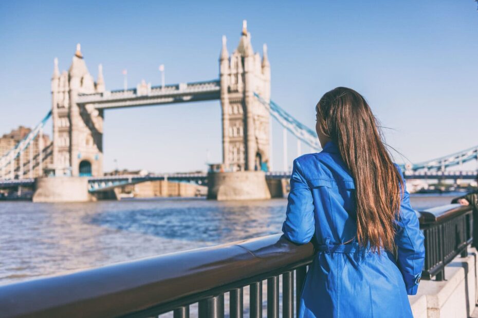 A person in a blue coat stands by a railing, gazing at the Tower Bridge in London on a clear day.